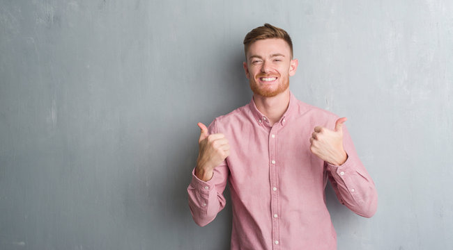 Young redhead man over grey grunge wall wearing pink shirt success sign doing positive gesture with hand, thumbs up smiling and happy. Looking at the camera with cheerful expression, winner gesture.