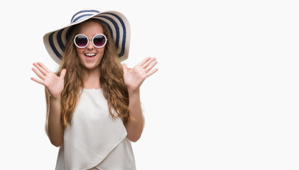 Young blonde woman wearing sunglasses and summer hat very happy and excited, winner expression celebrating victory screaming with big smile and raised hands