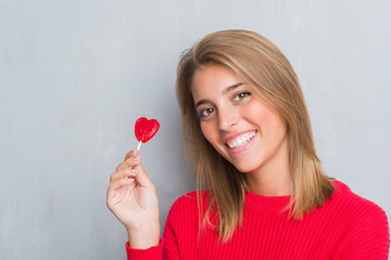 Beautiful young woman over grunge grey wall eating red heart lollipop candy with a happy face standing and smiling with a confident smile showing teeth