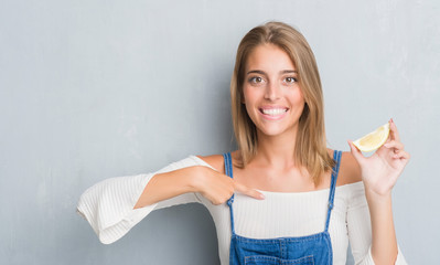 Beautiful young woman over grunge grey wall holding a lemon slice with surprise face pointing finger to himself