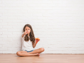 Young hispanic kid sitting on the floor over white brick wall eating pizza slice pointing with finger to the camera and to you, hand sign, positive and confident gesture from the front