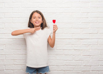 Young hispanic kid over white brick wall eating red heart lollipop candy very happy pointing with hand and finger