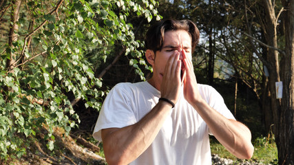 Young handsome man standing in nature, sneezing, ill from flu or cold
