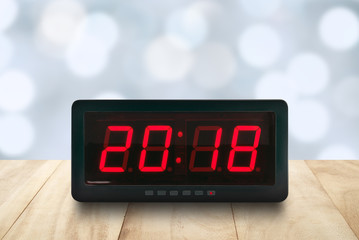 closeup red led light illuminated number 2018 on digital electric alarm clock face on brown wooden table top with defocused pastel blue and white Christmas lights bokeh background, time symbol concept