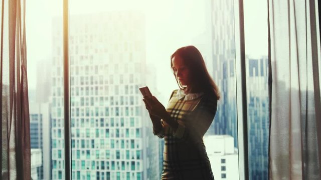 Beautiful young woman stands by the window uses smart phone on background of amazing sunset and blurred city in apartment. 3840x2160