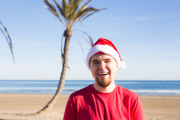 Obraz premium Christmas, people, holidays concept - Young man wearing a Santa hat smiling at the beach