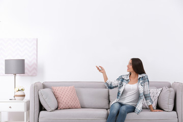 Obraz premium Young woman switching on air conditioner while sitting on sofa near white wall