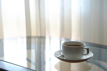 Ceramic cup of coffee with coaster and spoon on the glass table near the window behind curtain to outside the room