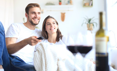 Young loving couple on sofa at home watching tv and laughing, drinking a glass of red wine.