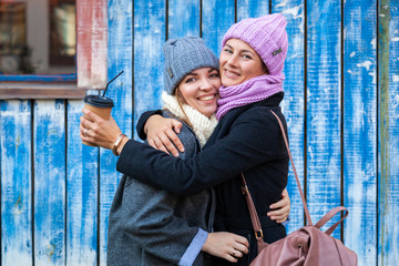 Obraz premium A young women friends in a stylish clothes hug, chat and drink coffee to go around blue wall. The concept of livestyle and outdoor recreation in autumn