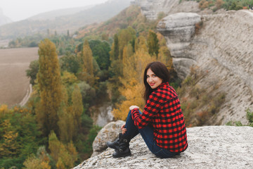 Naklejka premium Traveler woman wearing red plaid shirt and jeans sitting on mountain and looking at autumn forest. Hiking in autumn. Wanderlust concept. Cloudy weather, fall colors, autumn landscape.