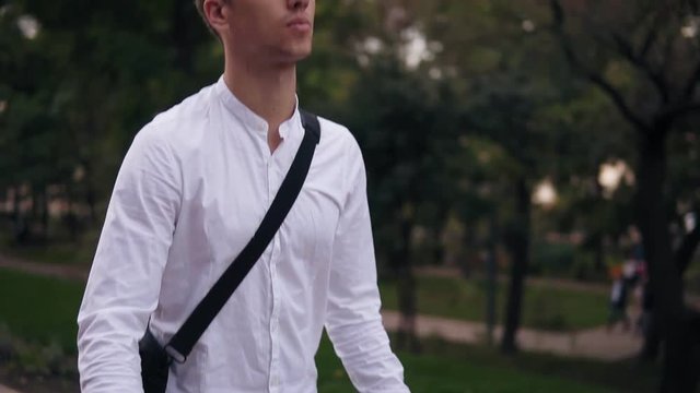 Caucasian young businessman walking with bicycle on the street in town. Rolling his trekking bike while walking by park with laptop case on shoulder. Slow motion