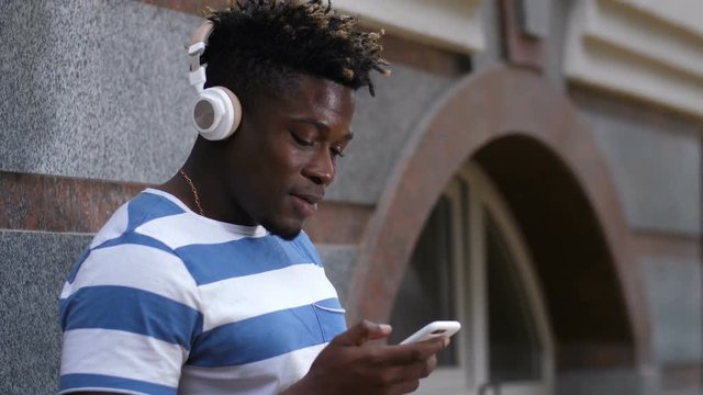 Good looking black man in t-shirt holding cellphone and listening to favorite funky music with bluetooth headphones on city street. Portrait of african american guy with smart phone enjoying music.