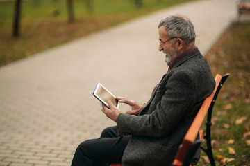 Obraz premium Grandpa uses a tablet and sits in the park on the bench. He look in to the screen