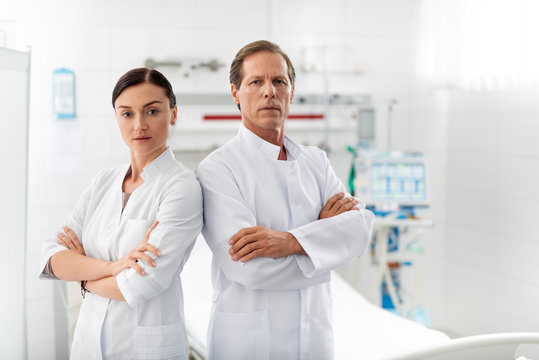 Reliable partners. Waist up portrait of middle aged doctor standing side by side with young nurse in white lab coat. They looking at camera with serious expression