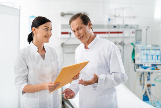 Waist up portrait of middle aged surgeon discussing patient illness history with colleague. Young lady looking at man with smile while holding documents