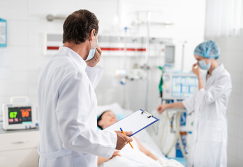 Back view portrait of surgeon in white lab coat holding clipboard and pen while touching protective mask on his face. Female medical worker talking with sick young lady on blurred background