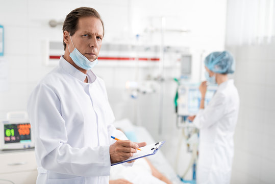 Waist up portrait of handsome surgeon with protective mask on chin noting information about patient while looking at camera with serious expression. Nurse near breathing machine on blurred background