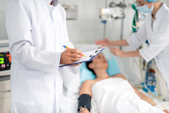 Cropped portrait of male therapist in white lab coat writing down information about condition of young lady on clipboard. Female assistant doing artificial inhalation on blurred background