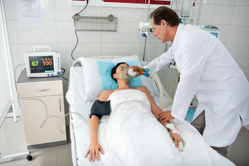 Young woman sleeping in hospital bed and getting mechanical ventilation. Middle aged man in white lab coat using respiratory equipment and touching lady arm
