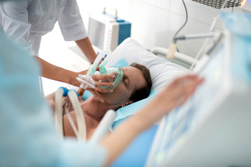 Close up of unconscious middle aged man getting mechanical ventilation in hospital and female hands using respiratory equipment