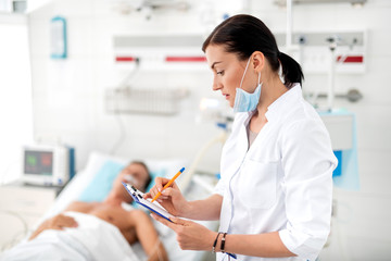 Side view portrait of female medical worker in white lab coat noting data about condition of sick man. Gentleman sleeping on hospital bed on blurred background