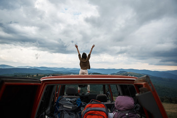 Full length of a young active female traveler standing on the car hood while rejoicing in a beautiful mountain scenery