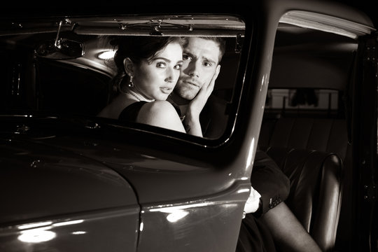 Good looking sexy couple, handsome man in suit, beautiful woman in red dress, are discovered embracing in vintage car