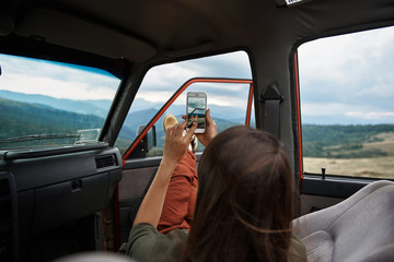 Rear view of a young delighted female traveler making shots of a beautiful scenery while travelling in the mountains by car