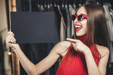 Pretty smiling woman wears red dress and sunglasses holding empty black card before shop clothes background, advertisement space