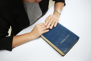 Woman with Bible praying, hands clasped together on her Bible on white table.