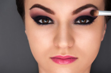 Close up shot of the young beautiful model with professional make up and pink lipstick and brush near her eye on the grey background