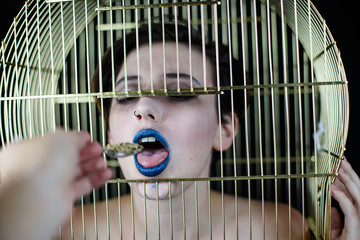 Conceptual shot of woman with blue lips and bird cage on head taking food from spoon 