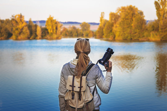 Woman holding a camera and looking for composition in autumn nature near to lake. 