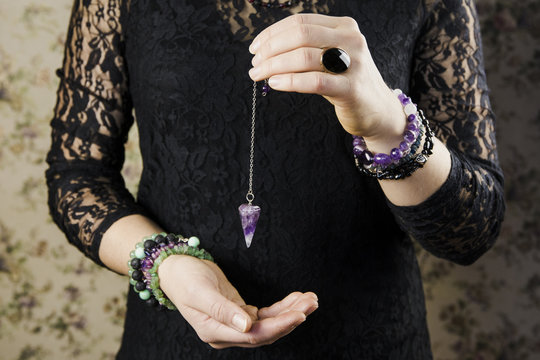 Close up of woman wearing black gothic clothing, hand holding and using amethyst crystal pendulum on silver chain fortune telling pendulum in hand. Mystical psychic dark witch concept. 