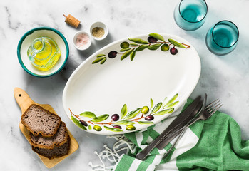 Empty handmade oval dish with olive tree branches on a white marble table. olive oil and cutlery. Mediterranean table setting