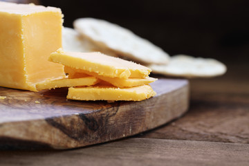 Block of cheddar cheese and slices over a rustic background.. Extreme shallow depth of field with selective focus on cheese. Water crackers in  blurred background.