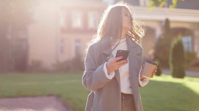 beautiful woman in brown coat goes near in the old city, communicates via smartphone (phablet). Steady cam shot, slow motion.autumn look