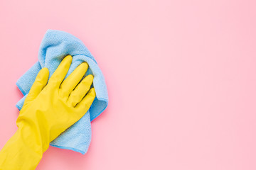 Employee hand in yellow rubber protective glove wiping pastel pink wall from dust with blue dry rag. General or regular cleanup. Commercial cleaning company. Copy space. Empty place for text or logo. - obrazy, fototapety, plakaty