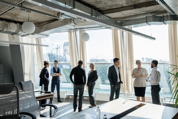 Group of modern business people standing at window with cityscape and discussing conference issues...