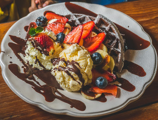 Chocolate Pancakes with banana, strawberry, blueberry, whipping cream and vanilla ice cream, icing sugar, brownies and chocolate sauce on wooden table.
