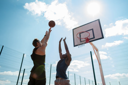 Low angle view of two men playing basketball and jumping reaching ball by hoop against sunlit blue sky, copy space