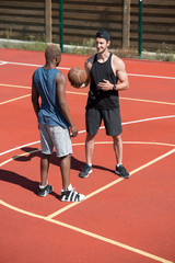 Full length portrait of two handsome  muscular men, one of them African, chatting standing on  basketball  court outdoors lit by sunlight, copy space