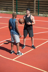 Obraz premium Full length portrait of two handsome muscular men, one of them African, chatting standing on basketball court outdoors lit by sunlight, copy space