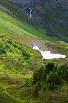 Narrow Hiking trail leading up apline green Alaskan Mountain side to distance waterfall