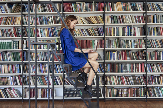 Beautiful young college student sitting on stairs in the library, working on laptop. Woman wearing blue dress, bookshelf on background with books.