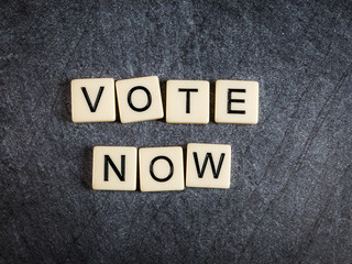 Letter tiles on black slate background spelling Vote Now