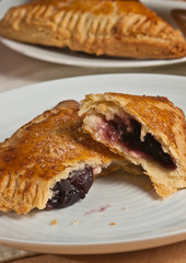 Front, top view, close distance of two freshly baked, homemade, cherry  cream turnovers and one ripped open to show cherry filling on a round white plate 