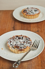 Top view, medium distance of  two, freshly baked, homemade cranberry raisin tarts on a round, white plate, covered with powdered sugar with an artisan fork on a wood serving board 