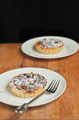 Top view, medium distance of  two, freshly baked, homemade cranberry raisin tarts on a round, white plate, covered with powdered sugar with an artisan fork on a wood serving board 